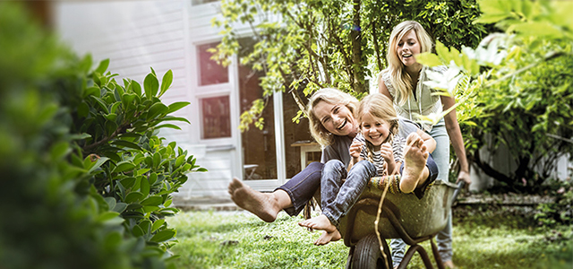Frau fährt ihre Mutter und kleine Tochter im Schubkarren im Garten herum – Symbolbild für Pflegerente