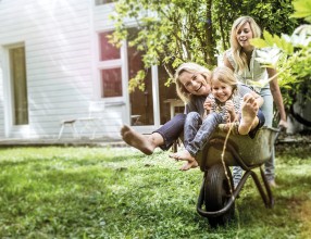 Frau fährt ihre Mutter und kleine Tochter im Schubkarren im Garten herum – Symbolbild für Pflegerente
